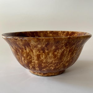 Pode incluir: Uma tigela de cerâmica envernizada em marrom e amarelo com um padrão manchado. A tigela é redonda e tem uma borda larga.