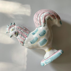 May include: A ceramic rooster figurine with a pink, blue, and yellow paint design. The rooster is standing on a pedestal base.