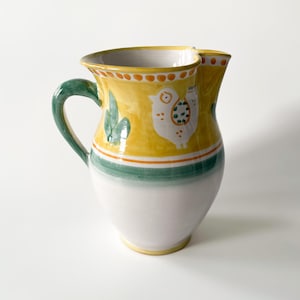 May include: A hand-painted ceramic pitcher with a yellow body, featuring a white chicken design and green cactus accents. The pitcher has a green handle, a white base, and a double spout design. The pitcher is approximately 8 inches tall.