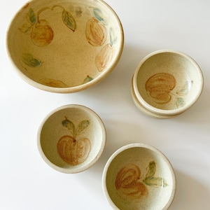 Puede incluir: Juego de cuencos de cerámica hechos a mano de varios tamaños. Los cuencos beige presentan diseños de manzanas marrones y verdes pintados a mano. El cuenco más grande tiene unos 25 cm de diámetro, y los cuencos más pequeños miden unos 10 cm.