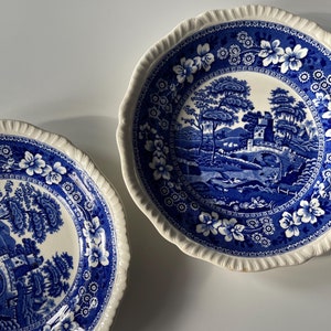 May include: Two blue and white porcelain plates featuring a floral design and a scene of a bridge over a river with trees and a building in the background.