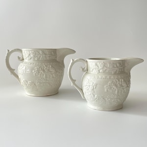 May include: Two white ceramic pitchers with embossed floral designs. The pitchers have a rounded body and a curved handle.
