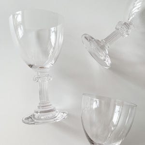 May include: Three clear, stemmed wine glasses with a classic design. The glasses have a wide bowl and a detailed, column-like stem. The glasses are arranged on a white surface, with one upright and two tilted.