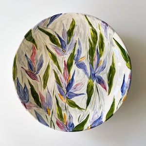 Op de afbeelding: Een grote, ronde keramische serveerschaal met een handgeschilderd bloemmotief. De schaal heeft een witte achtergrond met gestileerde blauwe, paarse, roze en groene bloemen en bladeren. Ideaal voor het serveren van salades of als decoratief stuk.