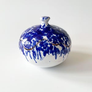 Op de afbeelding: Een ronde keramische container met deksel, witte basis en een opvallend blauw spetterpatroon. Het deksel heeft een kleine knop en een decoratieve golvende rand. Ideaal voor het opbergen van kleine items.
