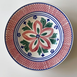 May include: A white ceramic bowl with a pink and green floral design. The bowl has a blue and red diamond pattern around the rim.