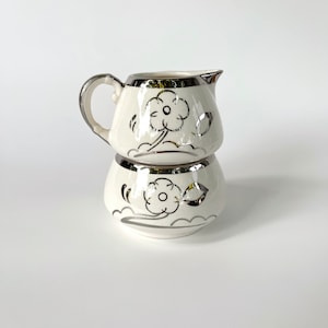 Puede incluir: Un juego de crema y azúcar de cerámica blanca con diseños florales plateados. La jarra tiene un asa y un pico. El azucarero tiene una tapa.