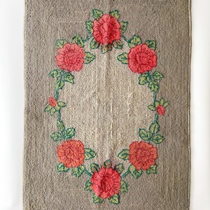 May include: Rectangular rug with a neutral gray background and a central floral design. The design features red roses with green leaves arranged in a wreath shape. The rug has a textured surface.