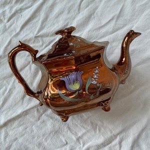 Peut inclure: Une théière de couleur cuivrée avec un motif floral. La théière a un bec courbé, une anse décorative et un couvercle avec un petit bouton. Le motif floral comprend des accents violets, verts et blancs. La théière est posée sur une surface blanche.