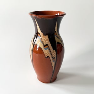 May include: A tall, terracotta ceramic vase with a flared rim. The vase has a dark brown upper section and a lighter terracotta lower section. Decorative vertical patterns in black, beige, and blue adorn the vase.