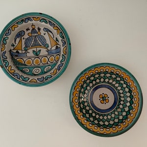 Könnte beinhalten: Zwei kleine Keramikschalen mit aufwendigen handbemalten Designs. Eine Schale zeigt ein blaues und gelbes Schiff mit einem grünen und gelben Rand. Die andere Schale hat ein grünes und gelbes geometrisches Muster mit einem blauen und weißen Zentrum.