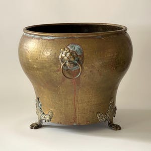 Puede incluir: Un macetero grande de latón antiguo con asas de cabeza de león y patas decorativas. El macetero tiene una pátina desgastada y envejecida y es perfecto para exhibir plantas o flores.