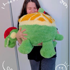 Puede incluir: Un gran cojín con forma de tortuga de felpa en tonos verdes, amarillos y rojos. El cojín tiene una superficie suave y texturizada y lo sostiene una persona. La palabra "HAPPY" está escrita encima de la cabeza de la persona.