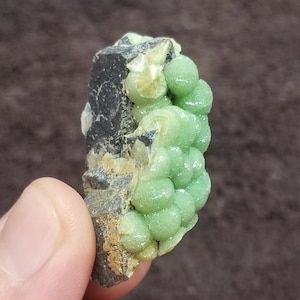 May include: A small green and black mineral specimen with a cluster of green crystals. The specimen is about 2 inches tall and is held in a hand.