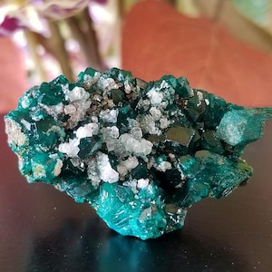 May include: A cluster of vibrant, teal-colored crystals with a sparkling, crystalline structure. The mineral formation has a rough, textured surface with patches of white and gray crystals. The background is blurred, featuring hints of pink and green.