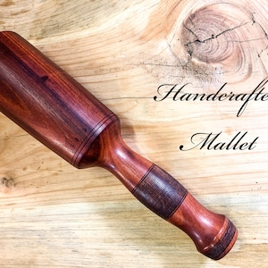 May include: Handcrafted mallet made of reddish-brown wood. The mallet has a cylindrical head and a handle with a textured grip. The text "Handcrafted Mallet" is written in cursive.