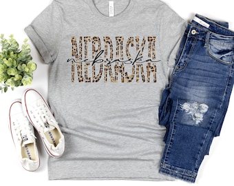 Peut inclure: Un t-shirt gris clair avec le mot "Nebraska" dans un motif léopard. Le mot "Nebraska" est écrit dans une police script. Le t-shirt est associé à un jean bleu et des baskets blanches.