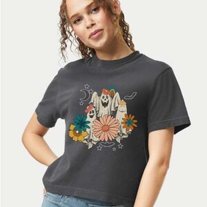 May include: Dark gray t-shirt featuring a retro-style graphic of three ghosts with floral accents. The ghosts have smiling faces and are surrounded by flowers, stars, a crescent moon, and a bat. The design has a vintage, whimsical aesthetic.