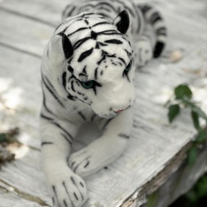 May include: A white plush tiger with black stripes, lying on a white wooden surface. The tiger has blue eyes and a pink nose.
