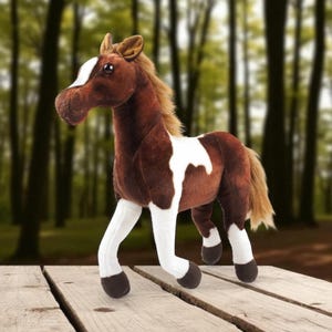 Puede incluir: Un caballo de peluche con pelaje marrón y blanco, de pie sobre una superficie de madera. El caballo tiene una mancha blanca en la cara y marcas blancas en el cuerpo y las patas. El fondo es una escena de bosque borrosa.