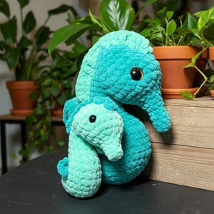 May include: Two crocheted seahorse plushies in shades of teal and turquoise. The larger seahorse has a textured body and dark brown eyes. The smaller seahorse is a lighter shade of teal. The plushies are on a black surface with potted plants in the background.