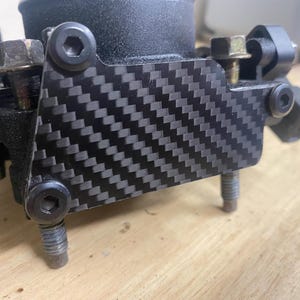 May include: Close-up of a black carbon fibre throttle body cover with six silver screws.