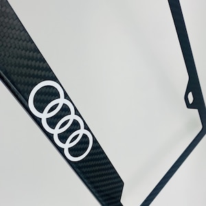 Puede incluir: Marco de matrícula de fibra de carbono negro con un logotipo Audi blanco.