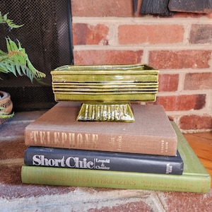 May include: A green ceramic planter with a square base and a rectangular top. The planter has a textured surface and is decorated with horizontal lines. The planter is sitting on top of three books.