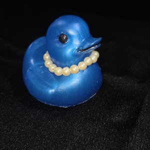 May include: A small, metallic blue rubber duck figurine featuring a smooth, glossy finish. The duck has a single black eye visible on the side of its head and is adorned with a necklace made of small, round, cream-coloured faux pearls.