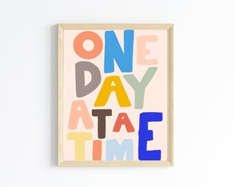 May include: A framed print with the text "ONE DAY AT A TIME" in colorful, stylized letters on a light pink background.