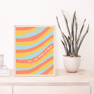 May include: A framed print featuring a colourful abstract design of wavy lines in yellow, orange, and blue. The text "All we have is now" is written in white on the print.