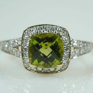 Op de afbeelding: Een ring met een kussengeslepen olijfgroene edelsteen in een halo van kleine witte steentjes. De ringband is gemaakt van metaal en is aan de zijkanten van de zetting bezet met extra kleine witte steentjes.