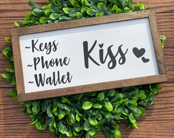 May include: A white wooden sign with a brown frame. The sign has black text that reads "Kiss" with a heart symbol. Below the word "Kiss" is a list of items: "Keys", "Phone", and "Wallet".