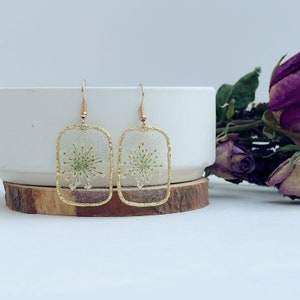 May include: A pair of gold rectangle earrings with a delicate white flower pressed inside each earring.