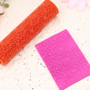Könnte beinhalten: Zwei Silikon-Nudelhölzer mit einem Blumenmuster. Das Nudelholz links ist orange und das Nudelholz rechts ist rosa.