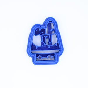 May include: A blue plastic cookie cutter shaped like two penguins standing on a platform. The penguins are wearing hats and scarves.