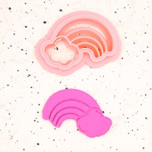 Puede incluir: Dos cortadores de galletas de arcoíris rosa, uno es rosa sólido y el otro es rosa translúcido. Los cortadores están sobre un fondo blanco con motas negras.