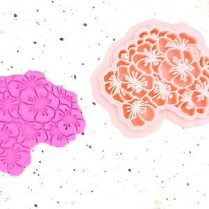 Può includere: Due formine per biscotti a forma di fiore. Una è rosa vibrante, che mostra il design floreale in rilievo. L'altra è una formina a strati nei toni del pesca e del bianco, con un motivo floreale dettagliato. Entrambe sono su una superficie bianca macchiata.