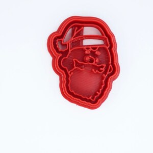 May include: Red plastic cookie cutter shaped like Santa Claus's face. The cutter has a detailed design of Santa's beard, hat, and eyes.