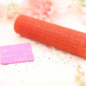 May include: A red silicone rolling pin with a geometric pattern. The rolling pin is on a white surface next to a smaller pink silicone square with the same pattern.