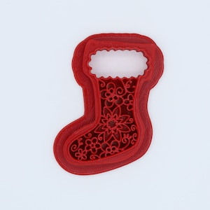 May include: Red Christmas stocking cookie cutter with a floral design. The cookie cutter has a detailed floral pattern within the stocking shape, perfect for holiday baking. The cutter is made of a durable material.