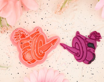 May include: Two pink and purple plastic cookie cutters shaped like a woman doing a yoga pose. The cutters are on a speckled pink background.