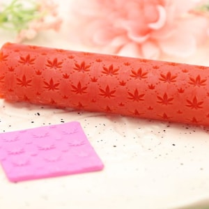 May include: A red rolling pin with a leaf pattern and a pink square rolling pin with a leaf pattern.