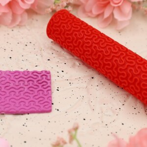 May include: A red and pink silicone rolling pin and a square silicone mold with a wavy pattern. Both tools are used for creating decorative designs on fondant or clay.