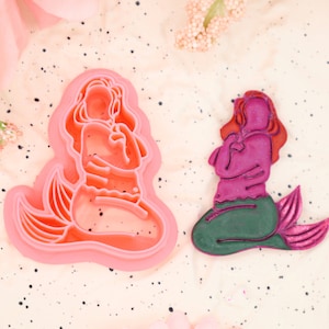 May include: Two mermaid cookie cutters, one pink and one green, with a detailed design. The pink cutter is on the left and the green cutter is on the right.