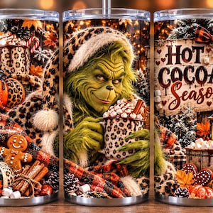 Può includere: Un bicchiere in acciaio inossidabile con un personaggio verde che indossa un cappello leopardato e tiene in mano una tazza di cacao. Il design include biscotti di pan di zenzero, zucche, pigne e la scritta "HOT COCOA SEASON" con un fiocco.