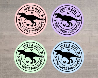 May include: Four round stickers with the text "JUST A GIRL WHO LOVES DINOSAURS". Each sticker features a black Tyrannosaurus Rex silhouette and small heart shapes. The stickers are in pastel colors: pink, purple, green, and blue.
