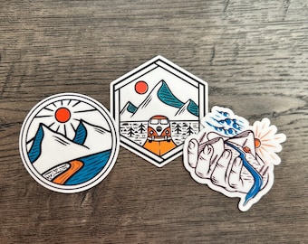 May include: Three colorful stickers with outdoor adventure themes. One is a circle with mountains, a river, and a sun. Another is a hexagon with a mountain scene and a VW bus. The third shows a hand holding a river and sun.