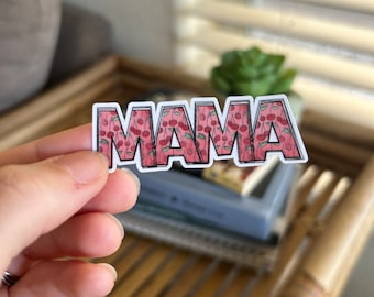 May include: A rectangular sticker with the word "MAMA" in a bold, outlined font. The letters are filled with a pattern of red cherries on a pink background. The sticker is held in a hand.