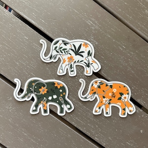 May include: Three elephant-shaped stickers featuring floral patterns. One sticker has a white background with orange flowers and green leaves. Another has a dark green background with orange and white flowers. The third has an orange background with white flowers.